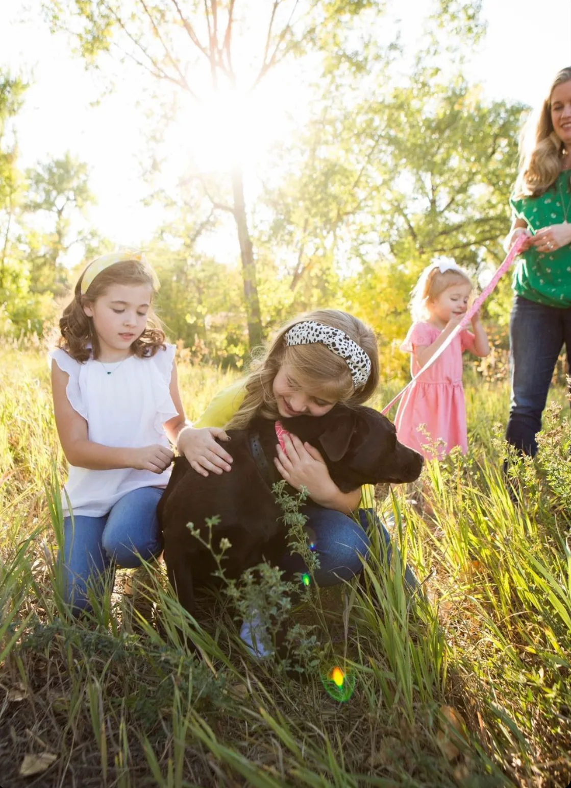 Two young girls hugging a black Labrador in a sunlit grassy field